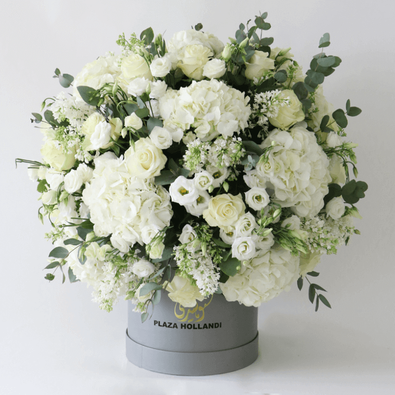 white flowers in a round design in a hat box white hydrangea, white roses, white wax flower and eucalyptus in a round design in a plaza hollandi hat box